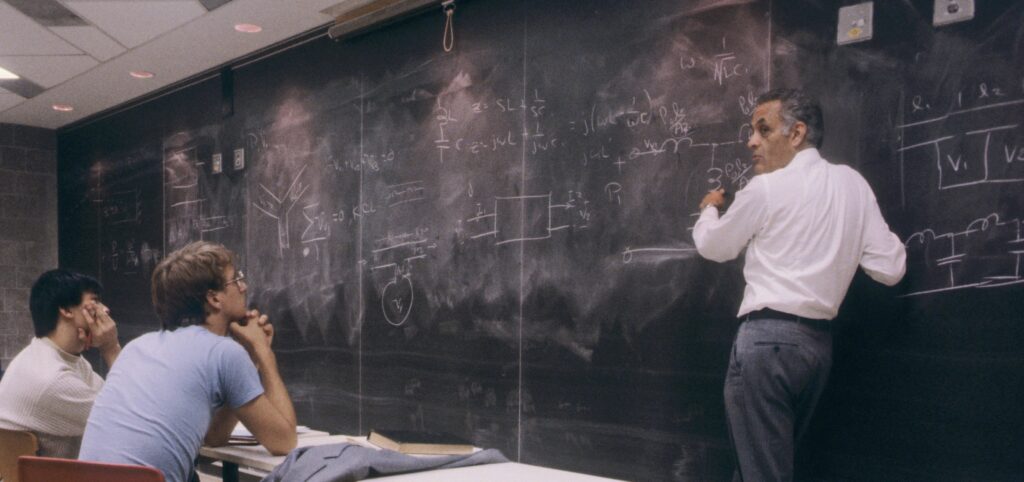 A professor writes equations on a chalkboard while students watch in a classroom.