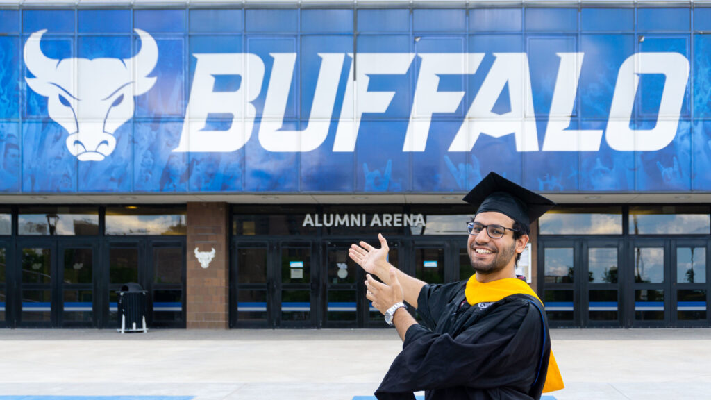 A man with glasses in university graduation robes stands in front of a giant "Buffalo" sign outside the University of Buffalo.