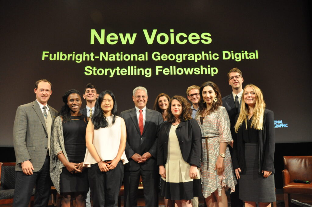 A group of Fulbright-National Geographic Fellows standing in front of a screen that reads: "New Voices - Fulbright-National Geographic Digital Storytelling Fellowship"