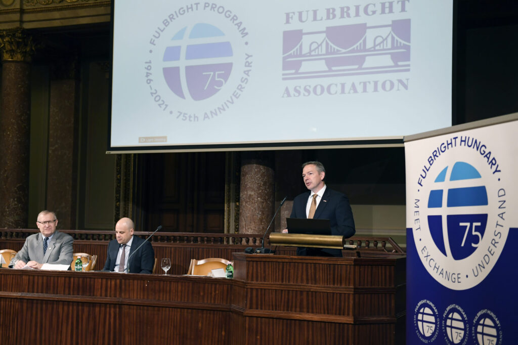 A man stands at a podium speaking and two others are seated beside him, sitting. In the background is a screen with the Hungarian Fulbright Association logo and the 75th Fulbright logo. On the standing man's other side is a pop-up banner with the Fulbright Hungary logo.