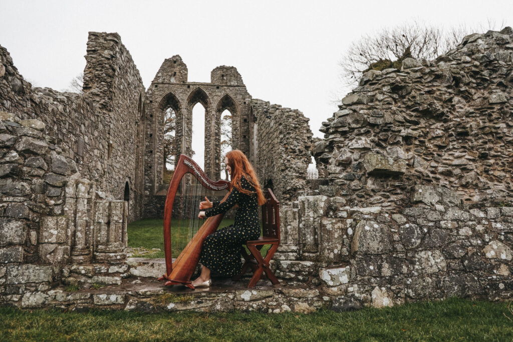 A woman with red hair and a long green dress plays harp in the outdoors among Irish ruins.