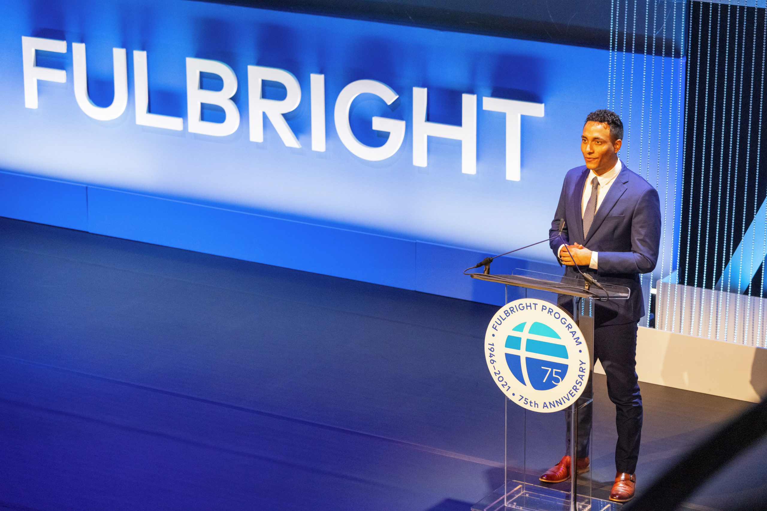 A person standing at a podium in a suit. The podium has a Fulbright logo on it and next to the person is "Fulbright" in big letters on the stage.