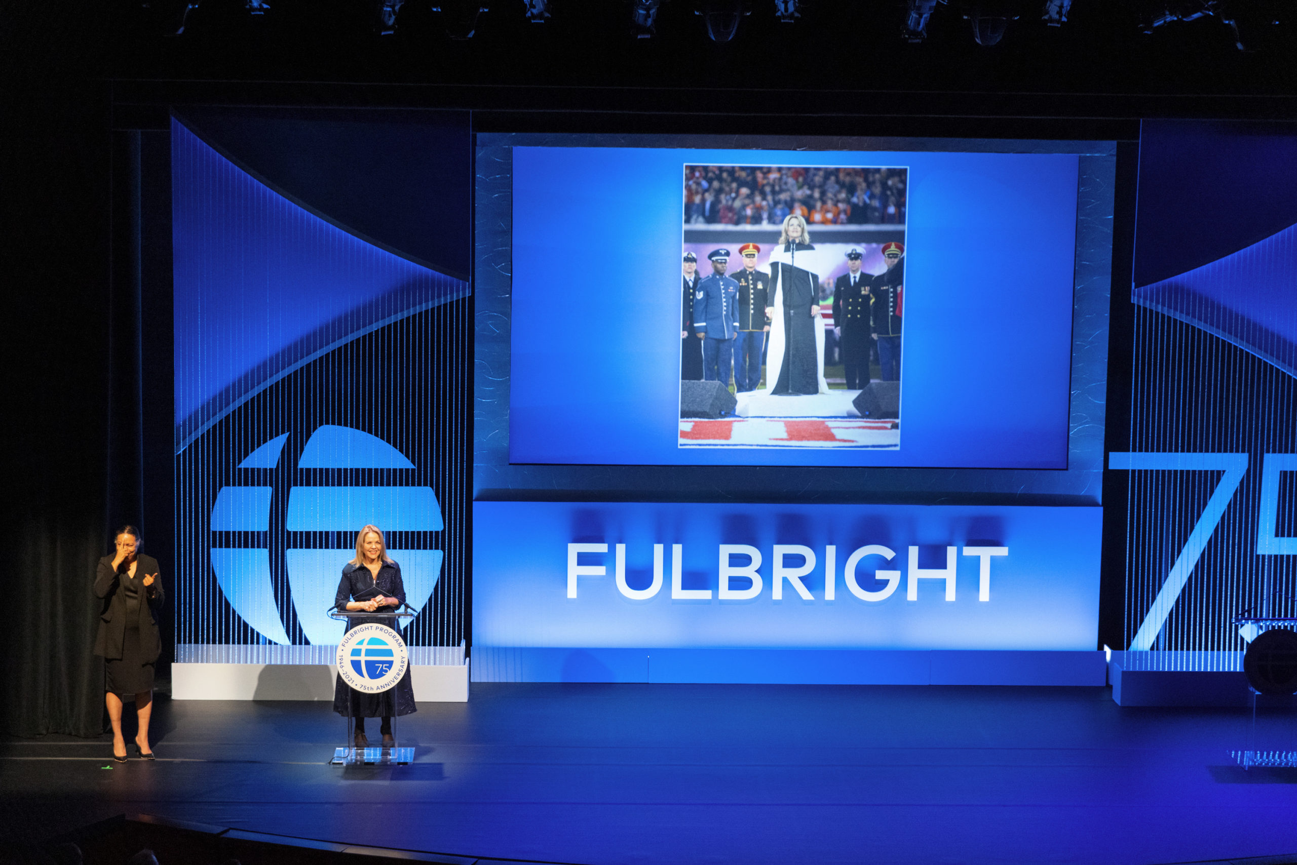 A person standing at a podium. The podium has a Fulbright logo on it and next to the person is "Fulbright" in big letters on the stage. A presentation is on a screen above them, with a photo of a person in graduation regalia.