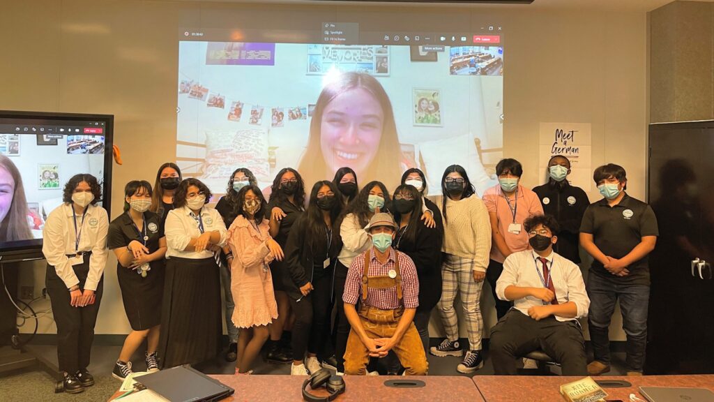 Photo of a group of students in masks standing in front of a wall; on the wall is a woman smiling from a video conference call.