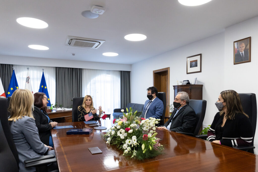 6 people sitting around a large wooden table in an office. Three are wearing masks. There is a large flower centerpiece on the table and some flags in the background.