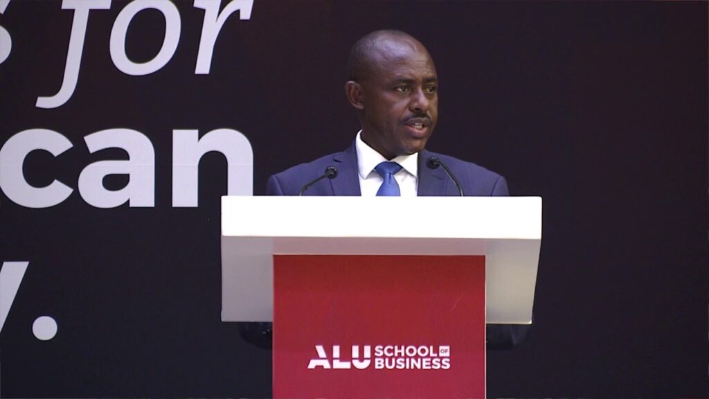 Dr. Eugene Mutimura speaking at a podium. The podium reads "ALU School of Business." He is addressing a group of graduates.