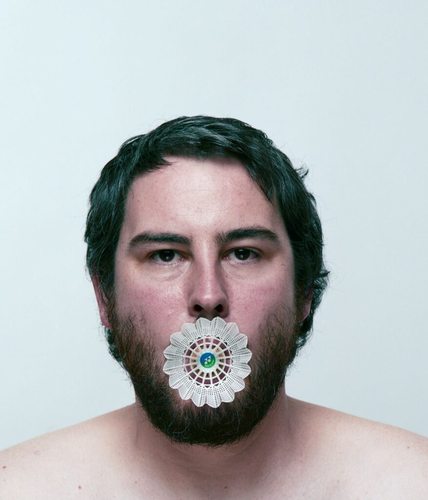 Artistic photograph of a person with dark hair and a dark beard with a badminton birdie in their mouth against a pale blue background.