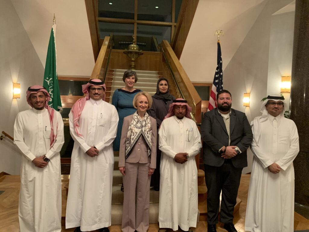 Six people standing in front, some in traditional Saudi Arabian wear. Two people stand in the back on a staircase.