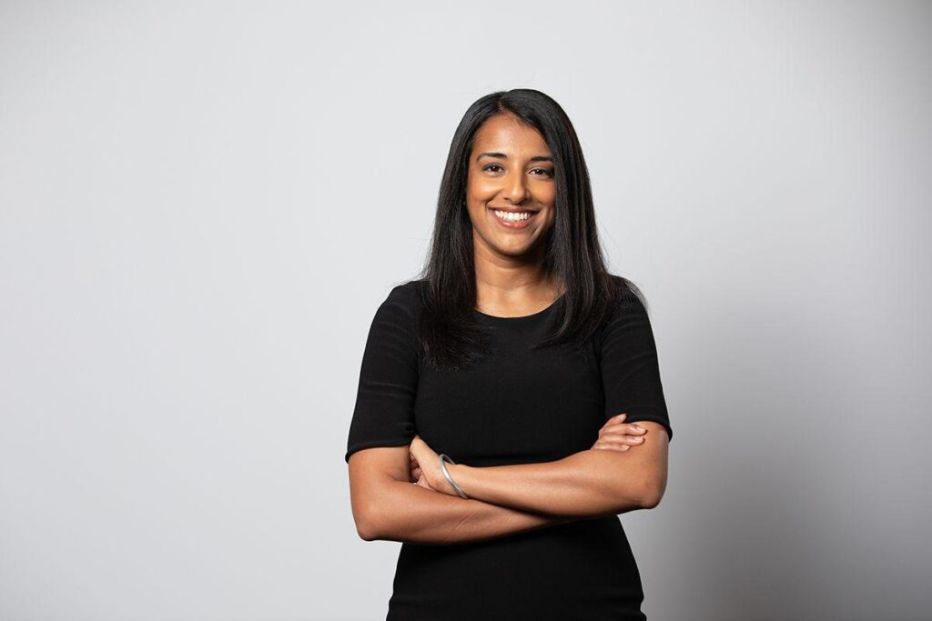 Person standing against wall, smiling and with arms crossed, in a black dress with quarter sleeves