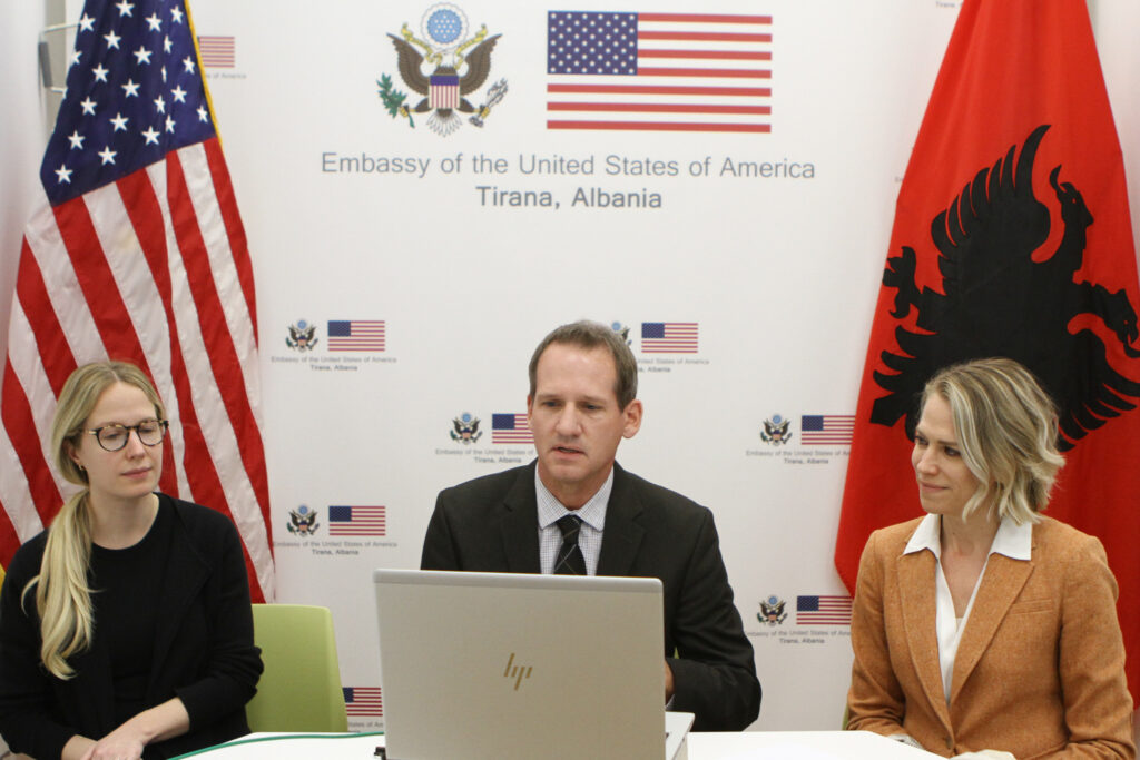 Three people sitting at a table with the U.S. flag and Albanian flag in the background