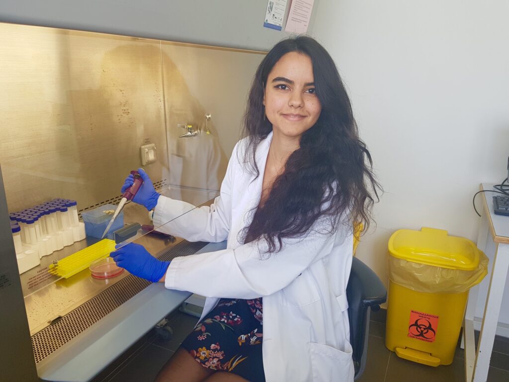 Person in a lab wearing a white lab coat and blue gloves, smiling at the camera as they hold a lab tool