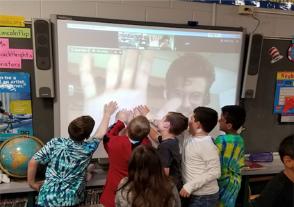 Large screen in classroom with Fulbrighter projected on it and young students in front waving at screen.