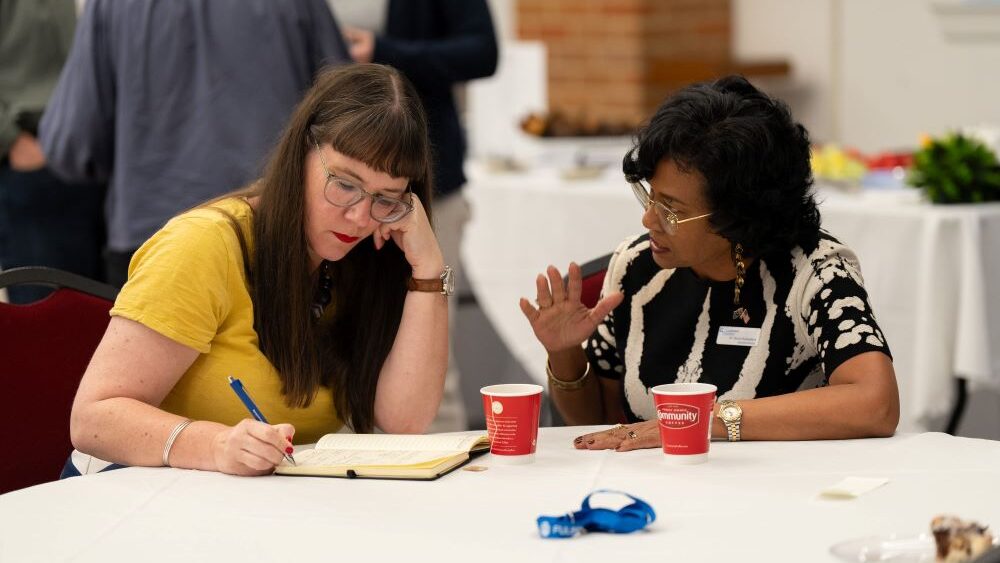 Two women sitting together at table, one writing on notepad and one talking. 