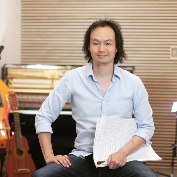 Christopher Tin sitting in front of musical instruments holding music sheets