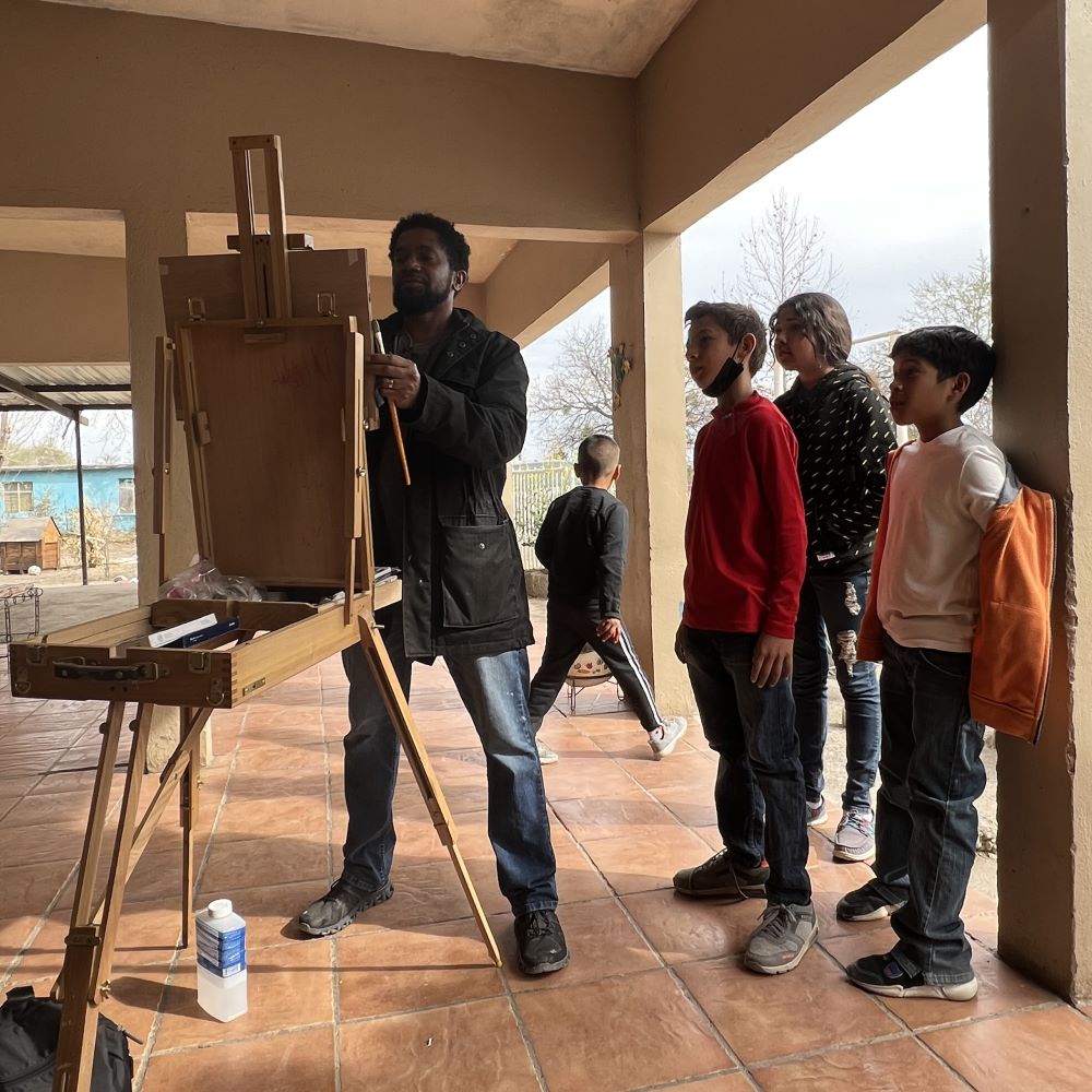 Sedrick Huckaby standing painting a easel with children standing by him
