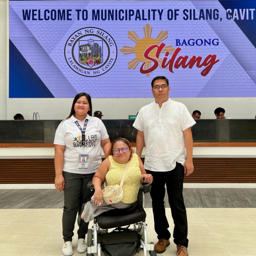 Olivia Mae Asuncion standing with two other people in front of large welcome sign projected on wall at the Cavite Institute