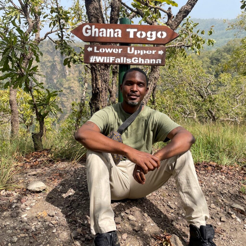 Karl Jackson sitting in front of tree and sign for Ghana Togo