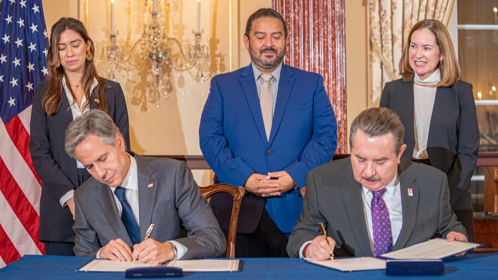 Secretary of State Antony J. Blinken sitting at table with gentleman signing documents, with three people standing behind them.