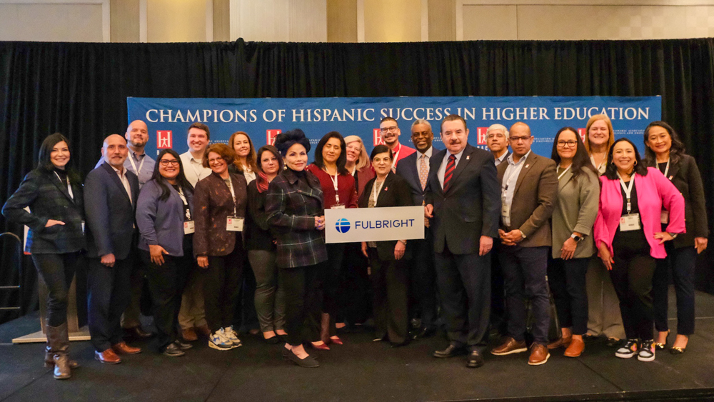 HSI leader representatives, Dr. Carmen Estrada-Schaye and HACU President Dr. Antonio R. Flores standing on stage at HACU conference holding Fulbright sign. 