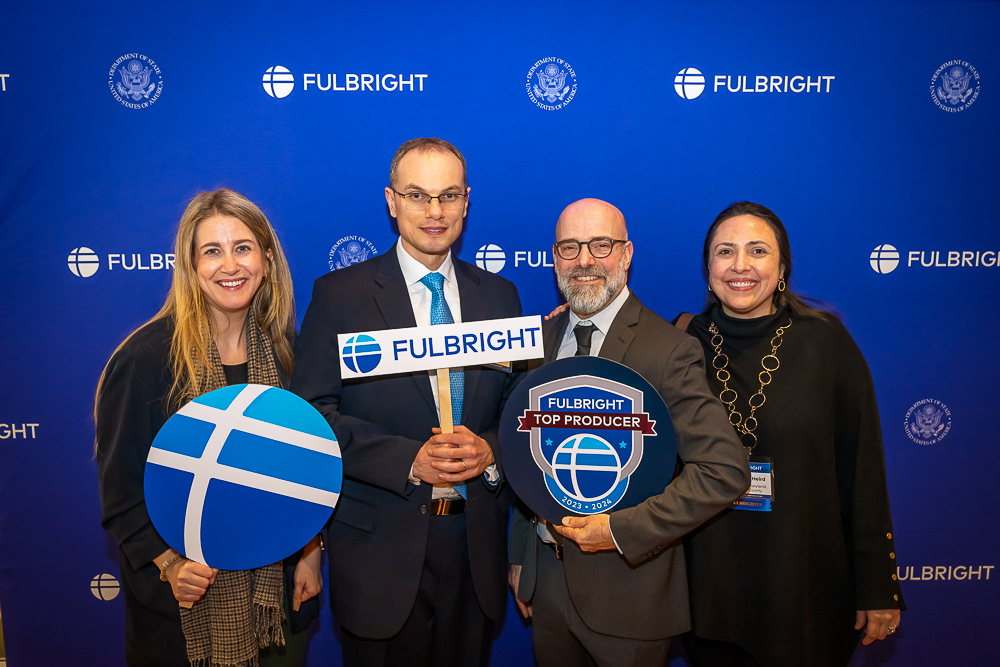 Four people posing with Fulbright signs against a blue background with Fulbright logos.