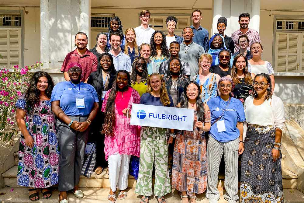 A diverse group of people holding a "Fulbright" banner, standing in front of a building with white columns and shutters in Senegal.