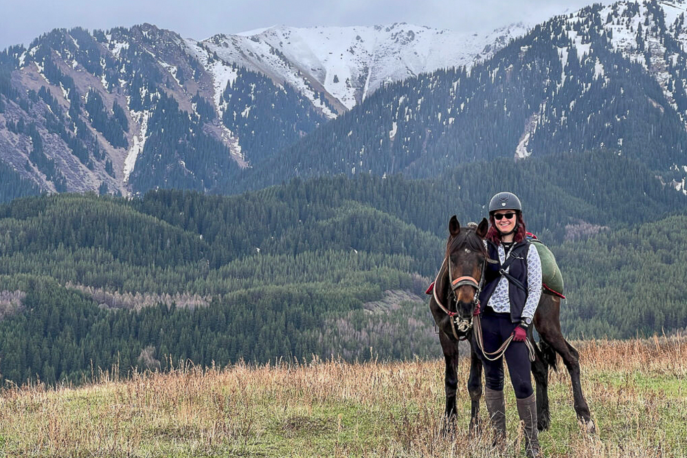 McKenna Wirth with a horse in a grassy field, with forested hills and snow-capped mountains in the background.