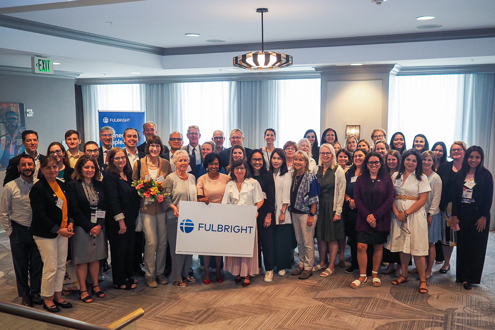 A group photo of approximately 40 Fulbright Commission EDs and staff standing in a conference room, with two women in front holding flowers and a Fulbright sign.