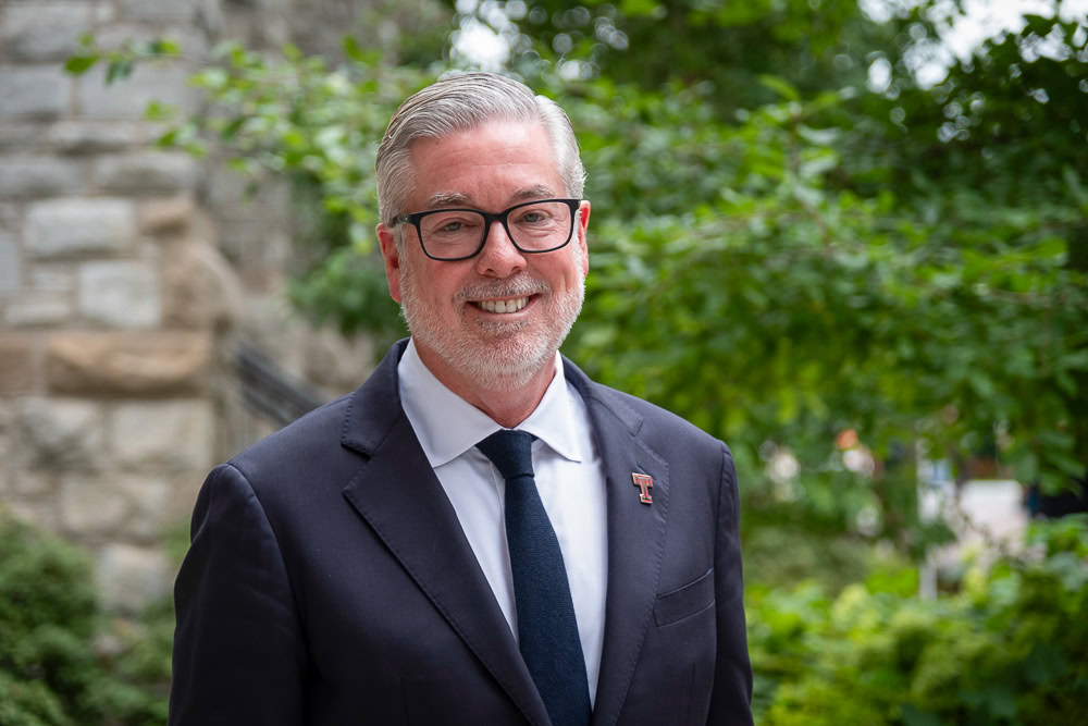John Fry in a suit with glasses standing in front of greenery.