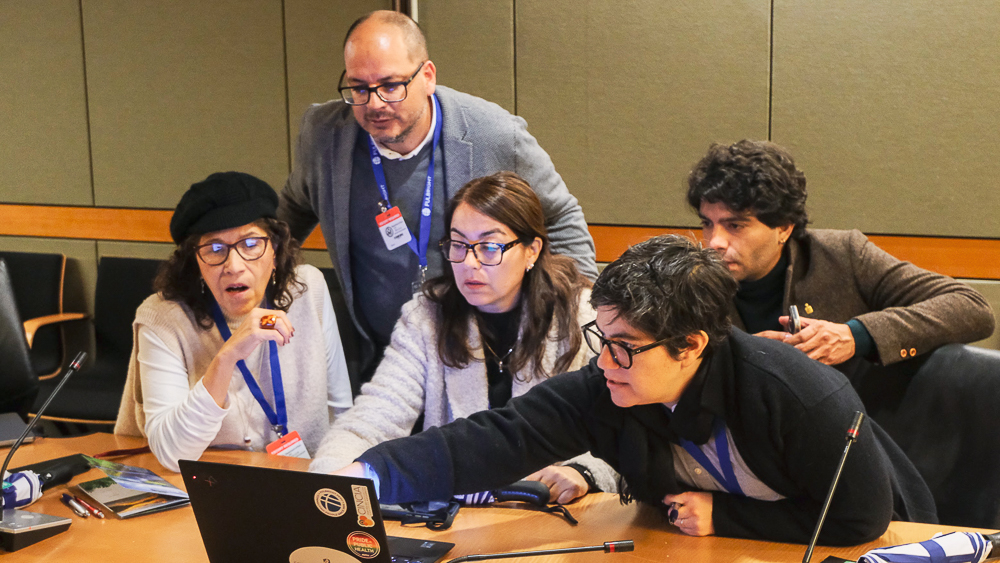 A group of five people gather around a laptop in a conference room, focusing intently on the screen.