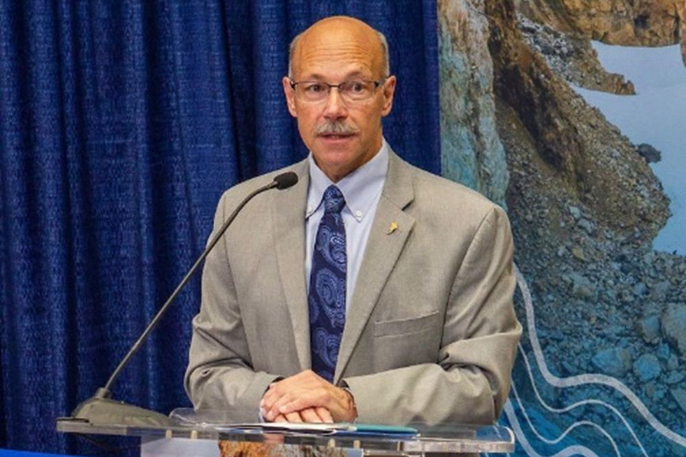 Sfraga in a gray suit speaks at a clear podium with a blue curtain background.