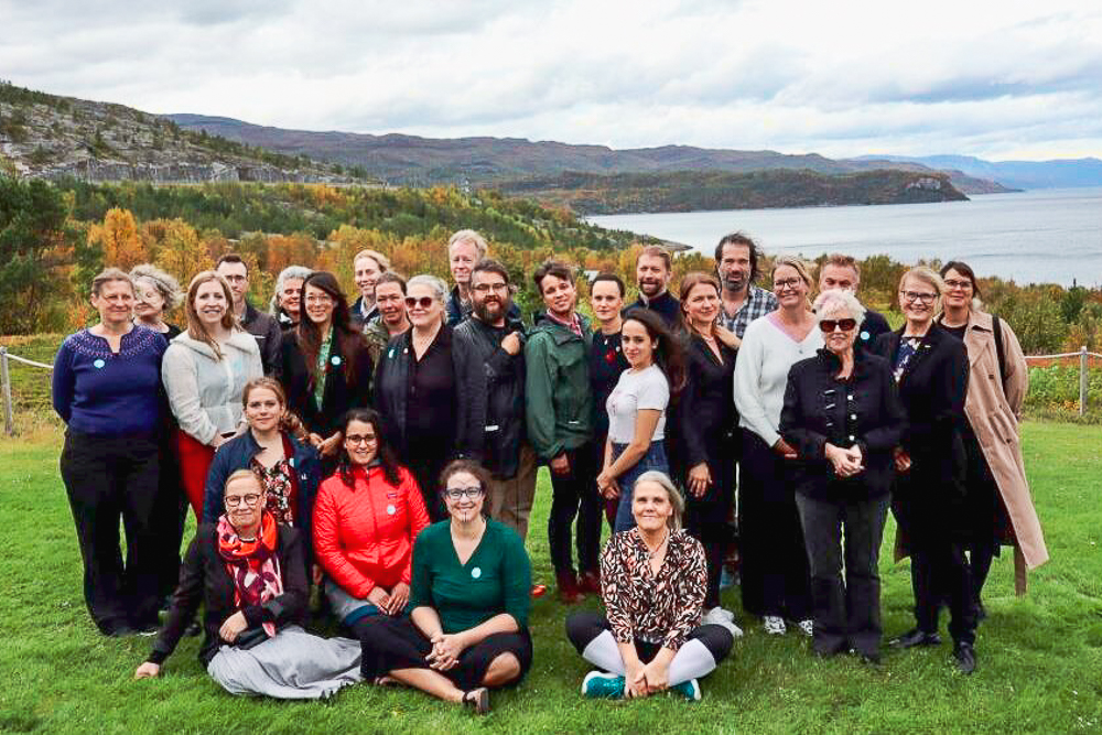 A group of people posed together on a grassy hill with a scenic backdrop of autumn trees and a body of water.