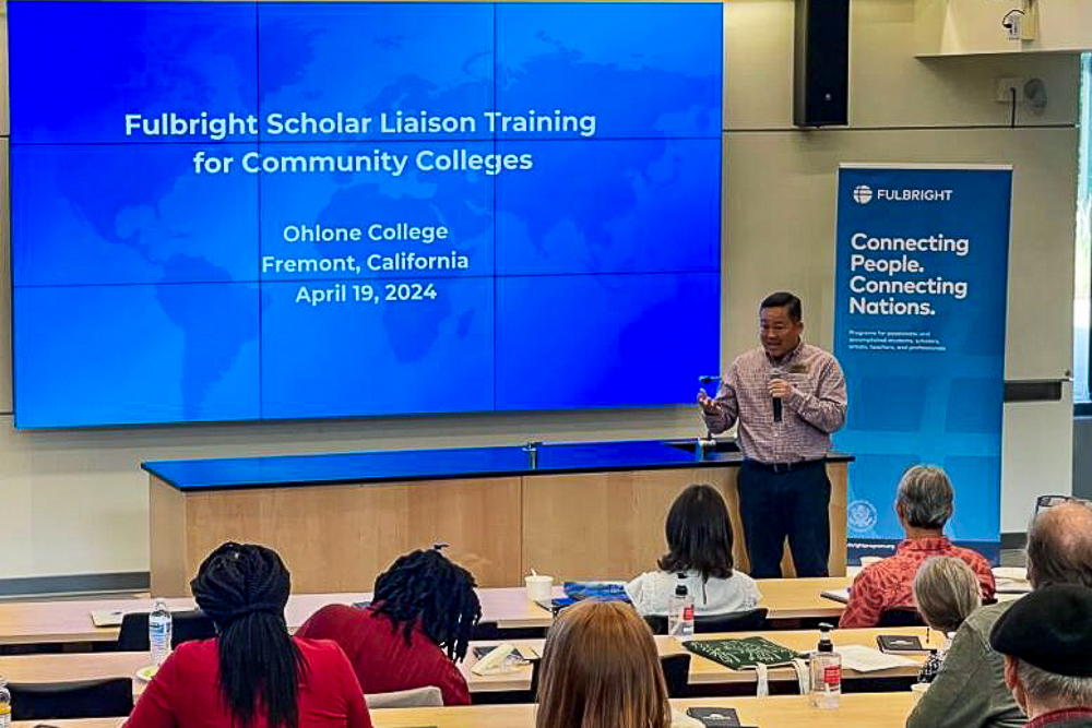 Charles presents at the Fulbright Scholar Liaison Training at Ohlone College with a large screen displaying event information.