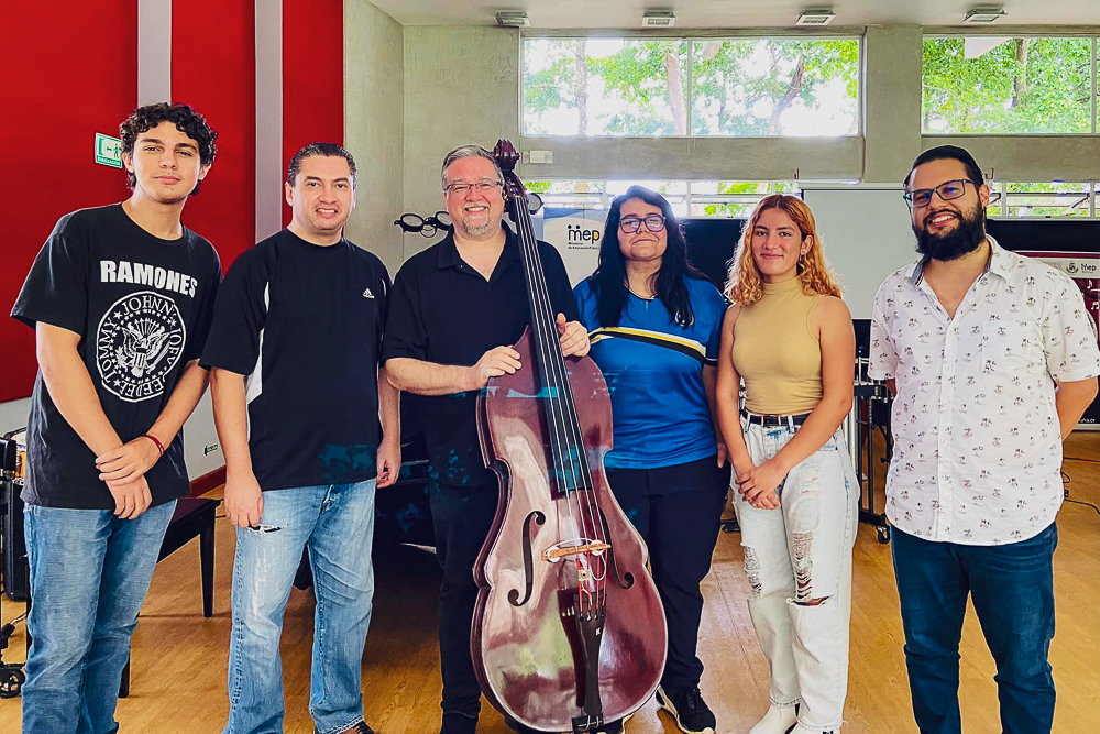 A group of six people stands indoors, with a man in the center holding a cello.