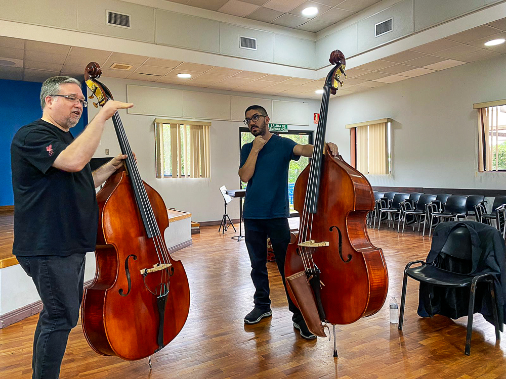 Two men with double basses in a room with hardwood flooring and chairs.