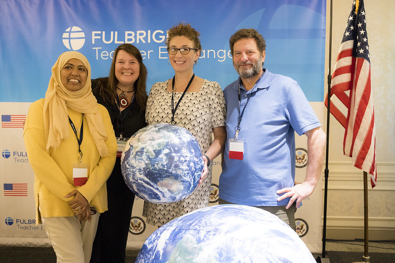 Four people standing with inflatable globes in front of a "FULBRIGHT Teacher Exchanges" backdrop and an American flag.