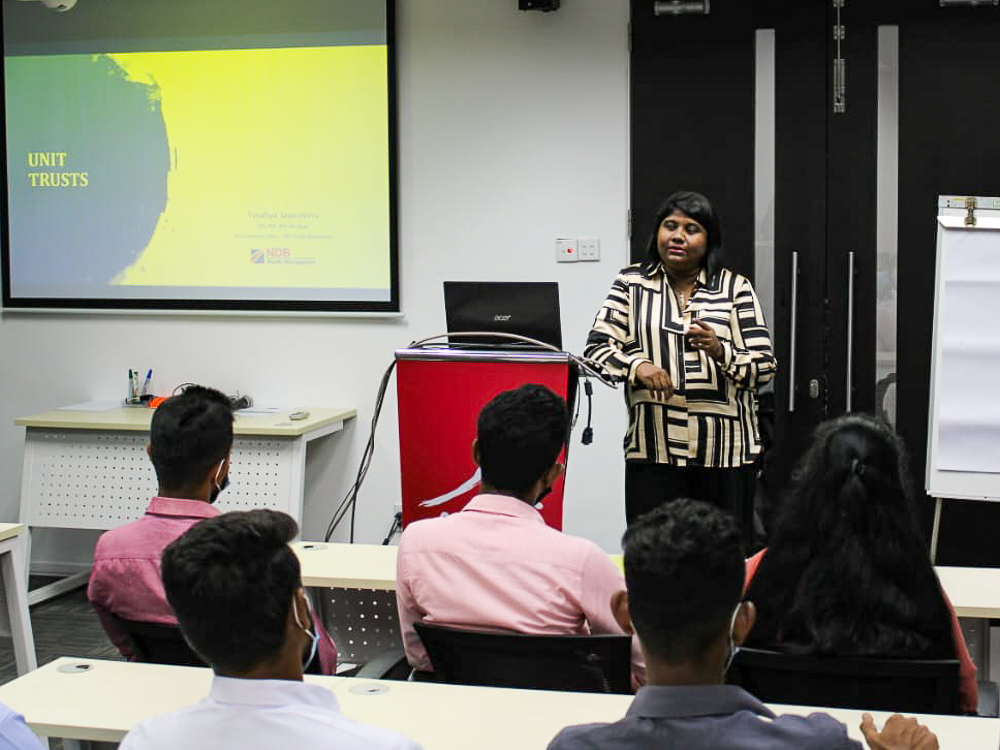 A woman presenting on "Unit Trusts" in a classroom setting with a projector screen and attendees.