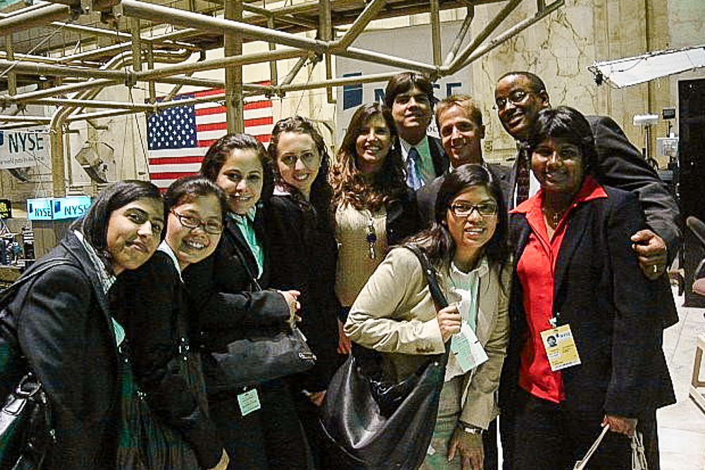 A group of ten people in formal attire smiling at the New York Stock Exchange, with an American flag and NYSE signs in the background.