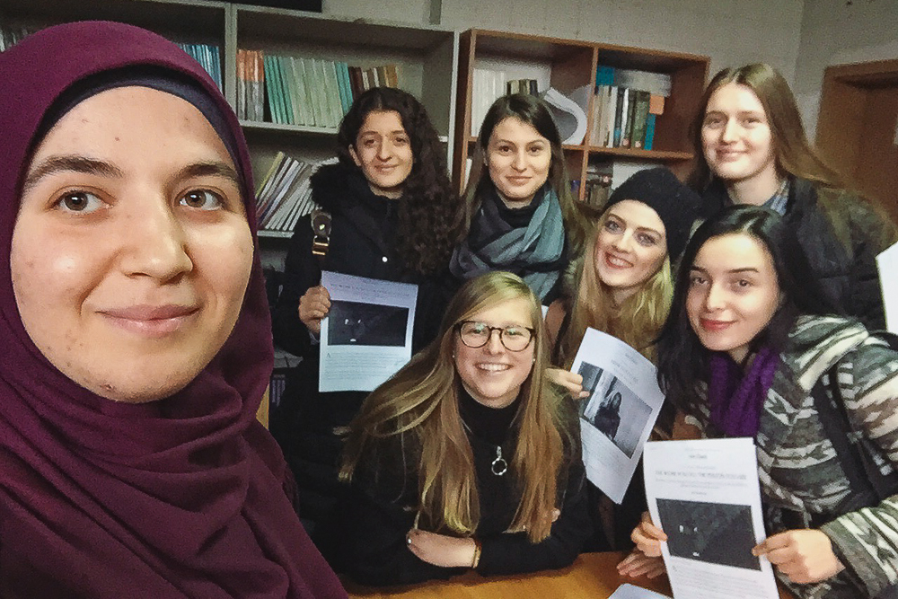 Seven women pose together in a room with bookshelves, holding papers and smiling.