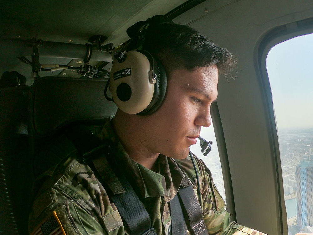 Benjamin Rich in military uniform with headphones looks out a helicopter window at a cityscape.