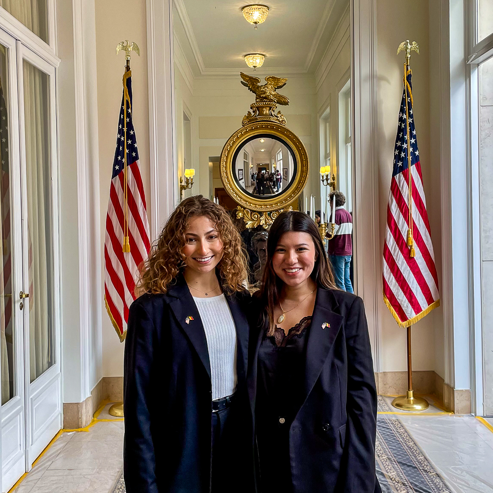 Two women in a hallway with American flags and an ornate mirror at the U.S. Embassy in Brussels