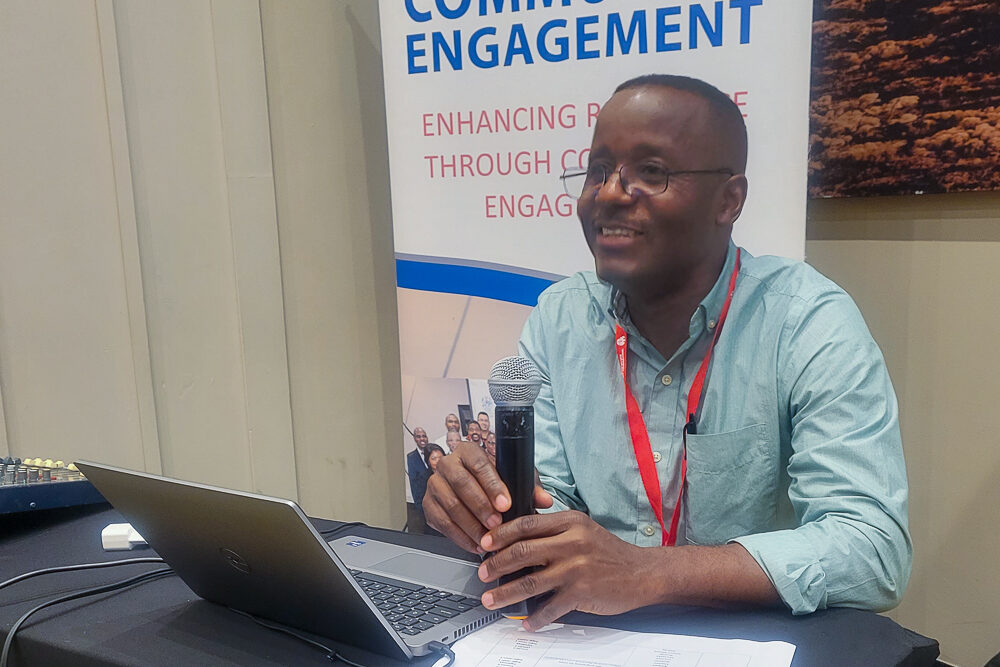 Wilson Majee speaking into a microphone at a conference table with a laptop.