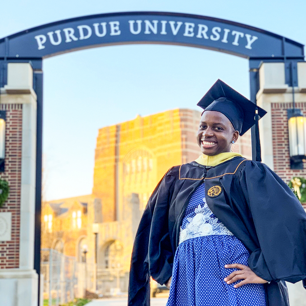 A graduate in cap and gown in front of a "Purdue University" archway.