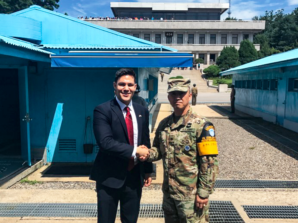 Two people shaking hands in front of blue buildings at the DMZ, one in a suit and the other in military uniform.