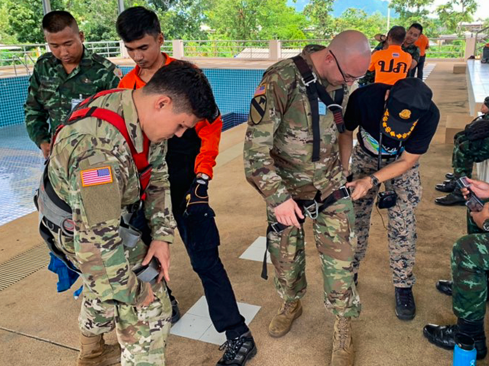 Group of individuals in military and civilian attire fitting harnesses near a pool.