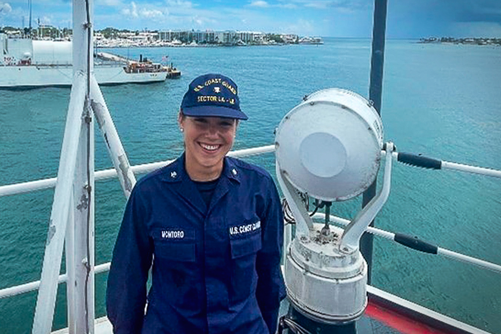 A person in a U.S. Coast Guard uniform stands on a ship next to a maritime light fixture, with water and a distant shoreline in the background.