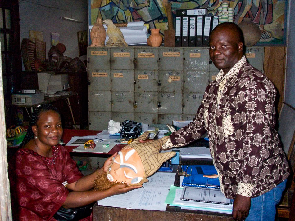 Two people in an office handling a wooden mask at a desk with background storage boxes and a mural.