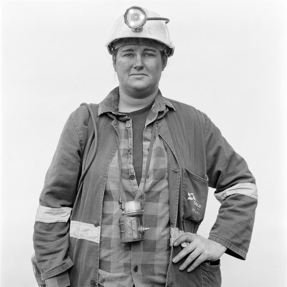 A person wearing a hard hat with a headlamp and work overcoat, standing with a neutral expression.