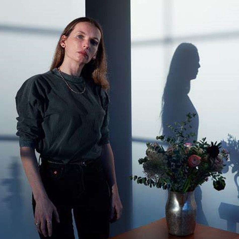 A woman stands indoors next to a table with a metallic vase of flowers, casting a shadow on the wall.