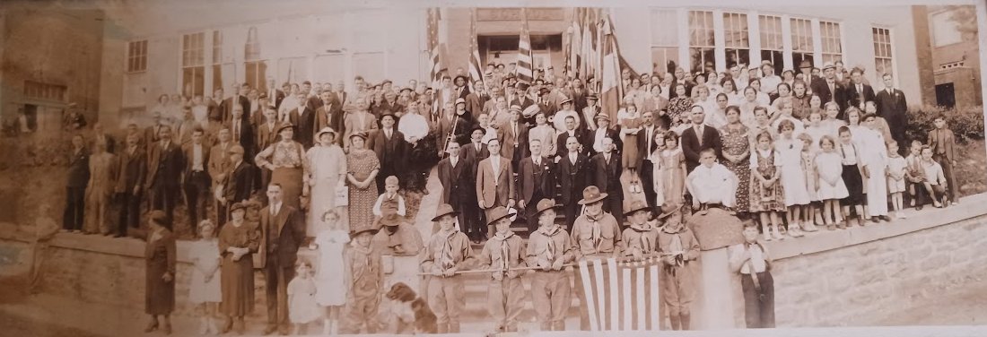 A sepia-toned historic group photo with many people gathered in front of a building.