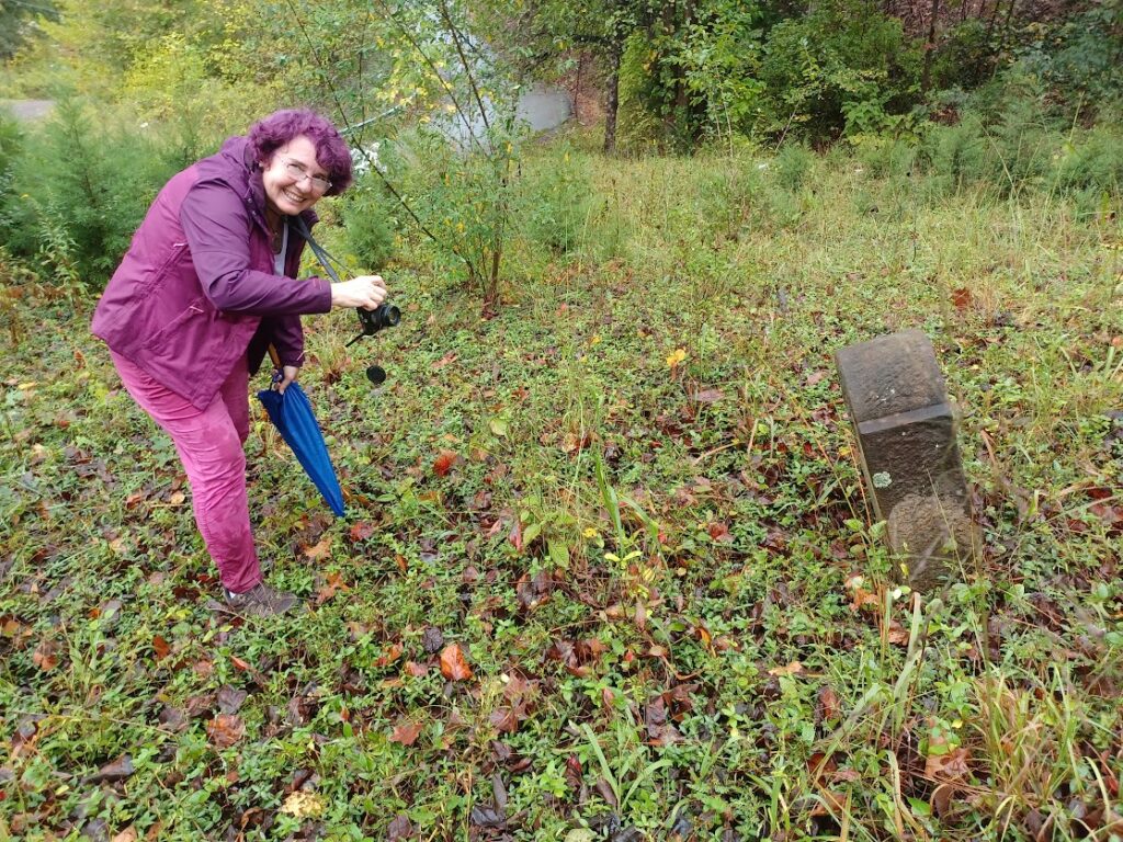 A person with a camera and blue umbrella beside a weathered tombstone in an overgrown grassy area.
